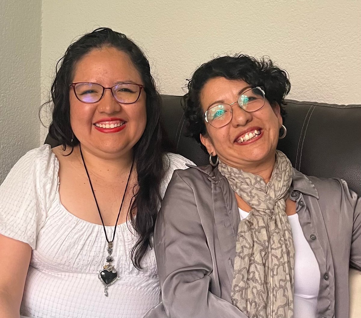 Two women smiling at camera, one wearing glasses and white shirt with lanyard, the other in gray blazer with patterned scarf