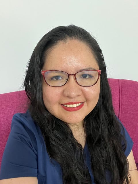 Portrait of a smiling woman with long dark hair, wearing pink-framed glasses and a dark blue shirt against a pink background