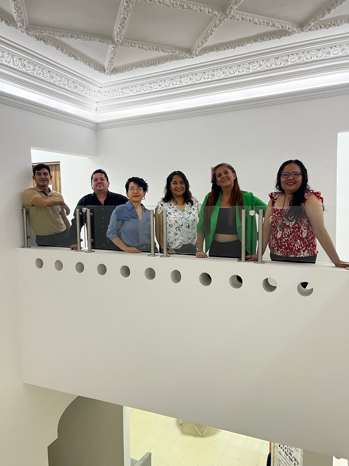 Six people standing together at an ornate white balcony railing with decorative ceiling details visible above them
