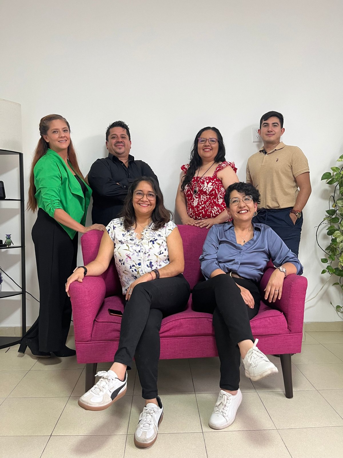 Group of six people posing together indoors, with two seated on a pink couch and four standing behind them
