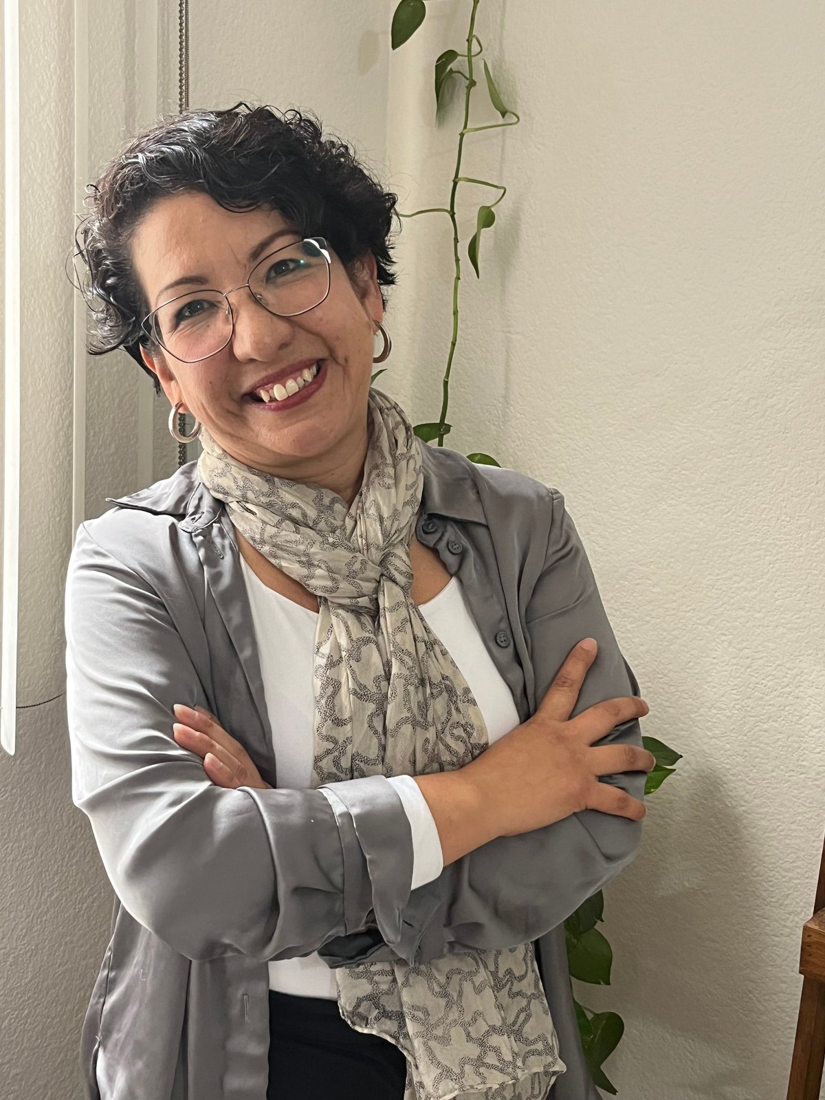 Portrait of a smiling woman with short dark hair and glasses, wearing a gray blazer and patterned scarf, posing with arms crossed
