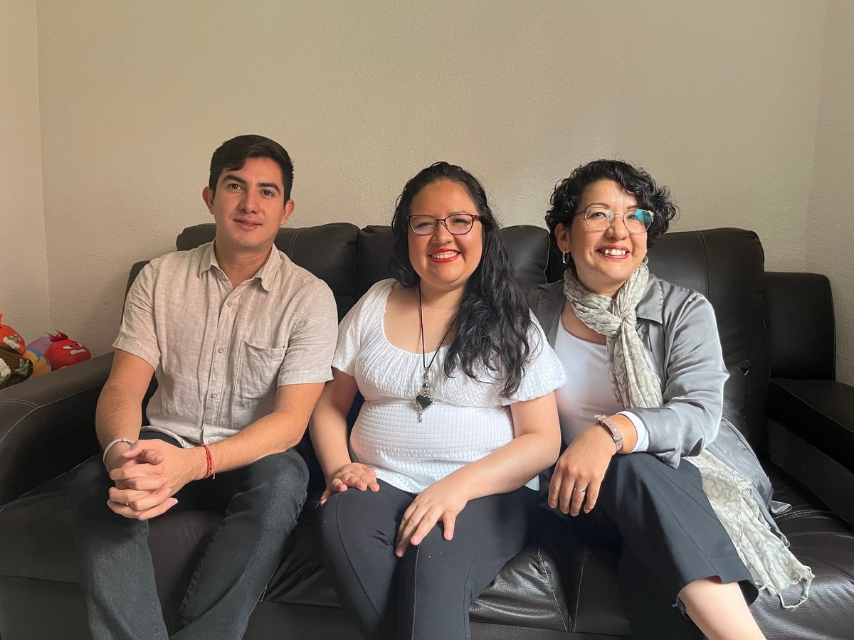 Three people sitting on a dark couch smiling at the camera in a casual indoor setting